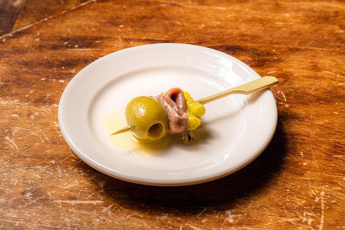 Product: A green olive, an anchovy, and a pickled guindilla pepper on a wooden toothpick on a small white dish, sitting on a wooden table - Cafe Ba-Ba-Reeba! in Lincoln Park - Chicago, IL Gluten Free Restaurants