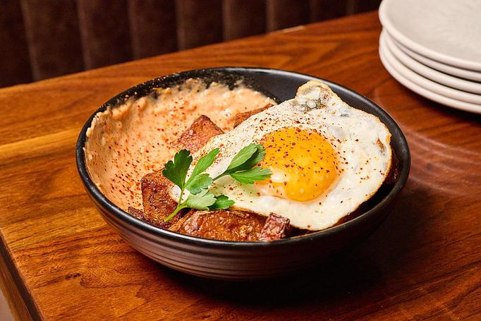 Product: A black dish with an orange sauce, a fried egg with a bright yellow center, golden potato pieces and a green garnish, on a wooden table - Cafe Ba-Ba-Reeba! in Lincoln Park - Chicago, IL Gluten Free Restaurants