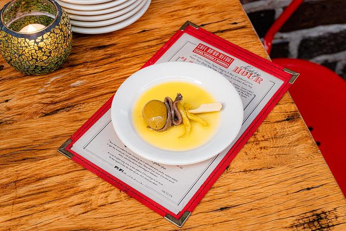 Product: A green olive, an anchovy, and a pickled guindilla pepper on a wooden toothpick in a pool of yellow olive oil in a small white dish, sitting on a Cafe Ba-Ba-Reeba! menu on a wooden table - Cafe Ba-Ba-Reeba! in Lincoln Park - Chicago, IL Gluten Free Restaurants