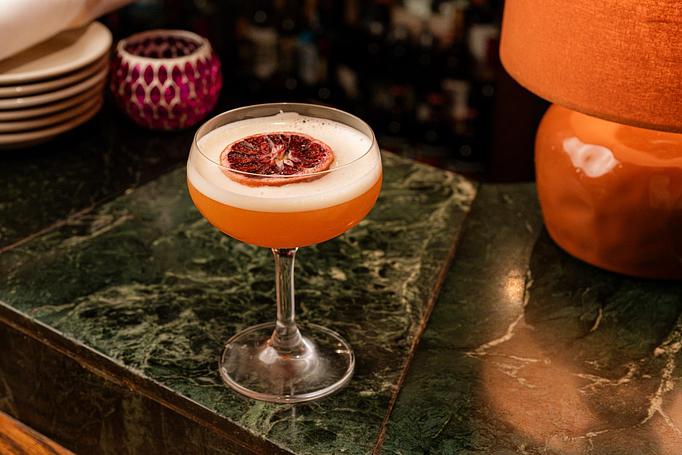 Product: A peach colored cocktail in a coupe garnished with a slice of preserved fruit, on a green marble bartop - Cafe Ba-Ba-Reeba! in Lincoln Park - Chicago, IL Gluten Free Restaurants