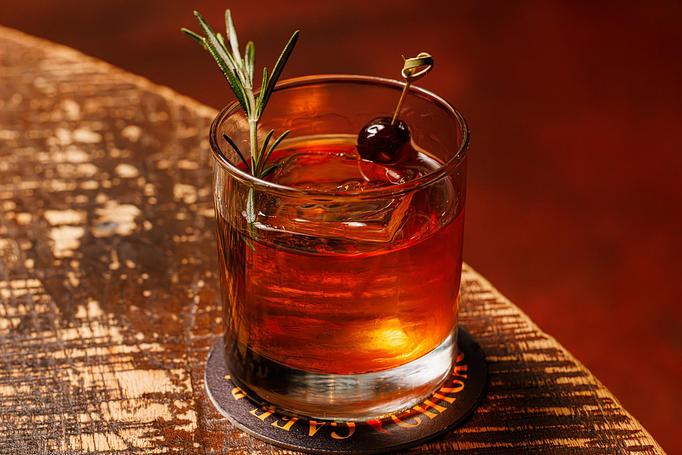 Product: A brown cocktail in a rocks glass with a large ice cube garnished with a sprig of rosemary and a berry, on a wooden table - Cafe Ba-Ba-Reeba! in Lincoln Park - Chicago, IL Gluten Free Restaurants