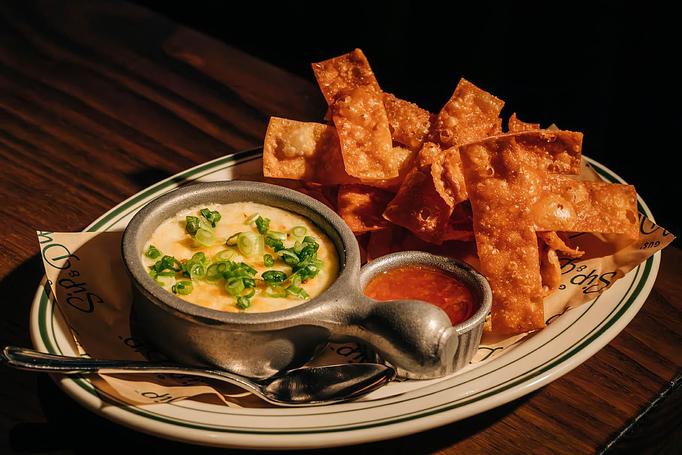 Product: A dish featuring a creamy dip topped with chopped green onions is served with crispy wonton chips. A small dish of red sauce accompanies the dish, set on a patterned plate with a napkin and spoon. - Gus' Sip and Dip in Chicago, IL Restaurants/Food & Dining