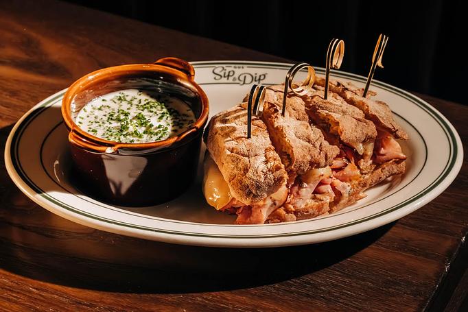 Product: A plate with a sliced sandwich filled with meat and cheese, secured with toothpicks, is presented. A small, brown bowl beside it contains a creamy sauce garnished with herbs. The plate is branded with "Gus' Sip & Dip." - Gus' Sip and Dip in Chicago, IL Restaurants/Food & Dining