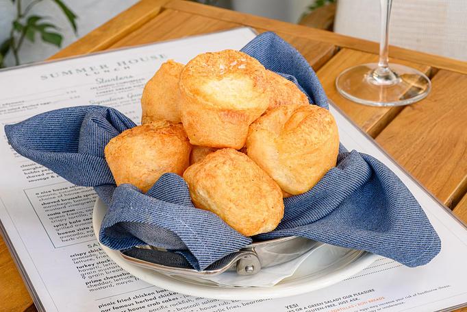 Product: A basket of golden, baked cheese puffs lined with a blue cloth napkin, placed on a plate atop a restaurant menu on a wooden table, with a wine glass partially visible in the background. - Summer House - Las Vegas in Las Vegas, NV Restaurants/Food & Dining