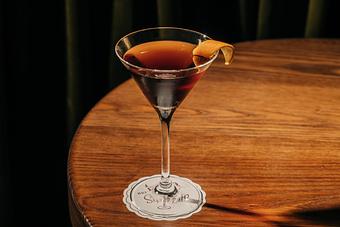 Product: A cocktail glass on a wooden table contains a dark liquid, garnished with a twist of orange peel. - Gus' Sip and Dip in Chicago, IL Restaurants/Food & Dining