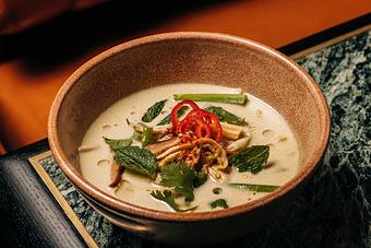 Product: A bowl of tom kha kai garnished with sliced red chili, fresh herbs, and mushroom pieces. Served in a brown bowl, set on a textured surface. - Crying Tiger in Chicago, IL Restaurants/Food & Dining