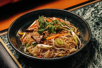 Product: A bowl of stir-fried glass noodles with vegetables, garnished with cilantro and fried garlic, served in a dark dish on a textured surface. - Crying Tiger in Chicago, IL Restaurants/Food & Dining