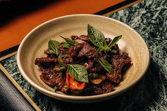 Product: A bowl of cumin lamb with basil leaves, sliced chili peppers, and herbs, placed on a patterned surface. The dish is vibrant with a mix of red and green colors. - Crying Tiger in Chicago, IL Restaurants/Food & Dining
