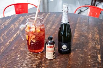 Product: A large clear pitcher of dark red liquid with ice, pieces of fruit, and wooden spoon in it, sitting next to a small bottle, and a bottle of Cava on a wooden table with red chairs in the background - Lil’ Ba-Ba-Reeba! in River North - Chicago, IL Bars & Grills