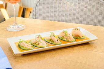 Product: Slices of hamachi are served on cucumber rounds with citrus sauce and microgreens, arranged on a long white plate on a wooden table, with a wine glass in the background. - Summer House - Las Vegas in Las Vegas, NV Restaurants/Food & Dining