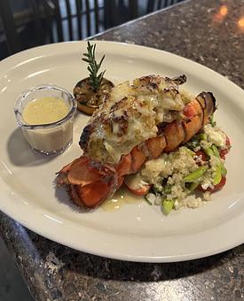 Product: Sweet lobster tail topped with crab cake, seated over quinoa mixed with feta, cherry tomato, asparagus, and spinach. Finished with a lemon-butter sauce. - The Bowman in Parkville, MD American Restaurants