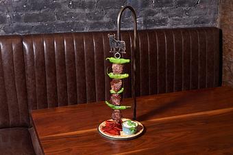 Product: An iron stand topped with the silhouette of a bull, with a kebab dangling off of it with pieces of beef, potatoes, and peppers, and a small blue and white patterned plate with small red peppers and a white sauce, all on a wooden table - Lil’ Ba-Ba-Reeba! in River North - Chicago, IL Bars & Grills