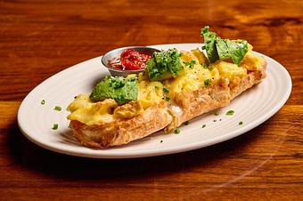 Product: A loaf of bread, halved, topped with bright yellow scrambled eggs, white cheese, and green avocado, served with a silver ramekin of red jam, on a wooden table - Lil’ Ba-Ba-Reeba! in River North - Chicago, IL Bars & Grills