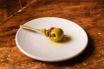 Product: A green olive, an anchovy, and a pickled guindilla pepper on a wooden toothpick on a small white dish, on a wooden table - Lil’ Ba-Ba-Reeba! in River North - Chicago, IL Bars & Grills