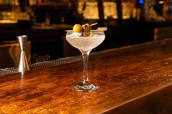 Product: A cloudy clear liquid in a clear coupe glass, garnished with a green olive, an anchovy, and a pepper, on a wooden bar top with a red stool in the background - Lil’ Ba-Ba-Reeba! in River North - Chicago, IL Bars & Grills