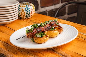 Product: A brown lamb skewer on golden potatoes topped with green herbs on a white oval plate on a wooden table - Lil’ Ba-Ba-Reeba! in River North - Chicago, IL Bars & Grills