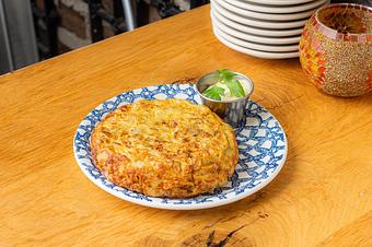 Product: A golden omelette with eggs and potatoes served with a white sauce in a silver ramekin garnished with green herbs on a blue and white patterned plate on a wooden table - Lil’ Ba-Ba-Reeba! in River North - Chicago, IL Bars & Grills