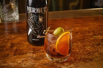 Product: Brown cocktail over ice in a short glass, garnished with an olive and an orange slice, sitting on a wooden table in front of a dark bottle of Vermut - Lil’ Ba-Ba-Reeba! in River North - Chicago, IL Bars & Grills