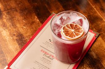 Product: A red cocktail in a clear glass with ice, topped with a preserved fruit slice, on a wooden tabletop - Lil’ Ba-Ba-Reeba! in River North - Chicago, IL Bars & Grills