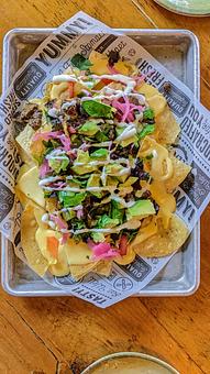 Product: Corn tortilla chips, cheese sauce, fresh pico de gallo, chopped greens, pickled jalapeño and onion, crema with your choice of brisket, pulled pork, or chicken (pictured here with Brisket and Avocado) - Longtable Beer Cafe in Downtown Middleton - Middleton, WI American Restaurants