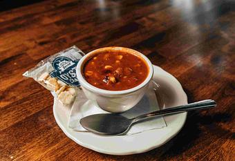 Product: a vegeterian chili served in a white cup on a white plate with a spoon and oyster crackers on a brown table - RJ Grunts in Lincoln Park - Chicago, IL American Restaurants