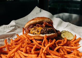 Product: A cheeseburger topped with bacon, served on a pile of sweet potato fries with sliced pickles, all on white parchment paper - RJ Grunts in Lincoln Park - Chicago, IL American Restaurants