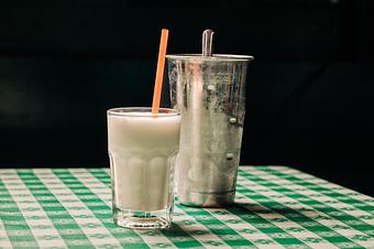 Product: A white milkshake served in a clear glass with a red straw, with a large metal cup in the background, set on a green-and-white checkered tablecloth - RJ Grunts in Lincoln Park - Chicago, IL American Restaurants