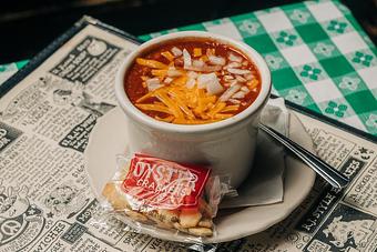 Product: A white bowl of chili topped with shredded cheese and chopped onions, served with oyster crackers, placed on top of an RJ Grunts menu on a table with a white and green checkered tablecloth - RJ Grunts in Lincoln Park - Chicago, IL American Restaurants