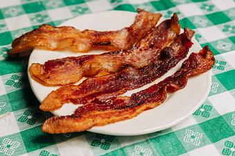 Product: Four pieces of crispy bacon arranged on a white plate atop a green checkered tablecloth - RJ Grunts in Lincoln Park - Chicago, IL American Restaurants