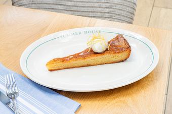 Product: A long, triangular slice of Warm Gooey Lemon Butter Cake. It features a dense, golden-yellow base with a slightly caramelized, glistening top layer. Garnished with lemon zest and whipped cream - Summer House - Las Vegas in Las Vegas, NV Restaurants/Food & Dining